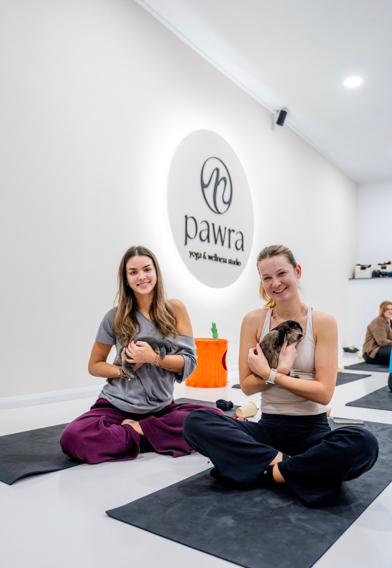 Two smiling women holding lop-eared bunnies on yoga mats at a Pawra bunny yoga class